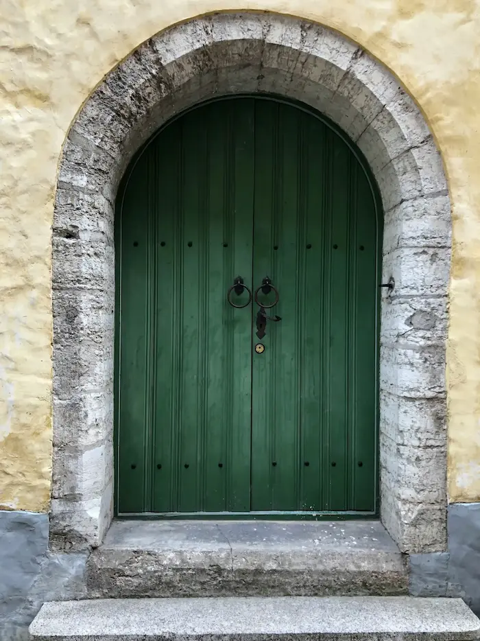 Une porte verte en bois avec des poignées en fer forgé, encadrée par un arc en pierre, avec un mur en pierre beige en arrière-plan.