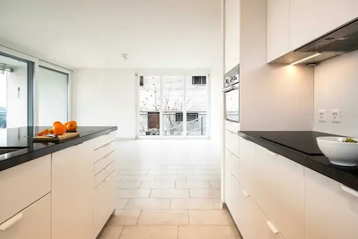 Une cuisine moderne avec des meubles blancs, un plan de travail noir, un évier, un four encastré, et une vue sur un balcon avec des arbres dénudés en arrière-plan.