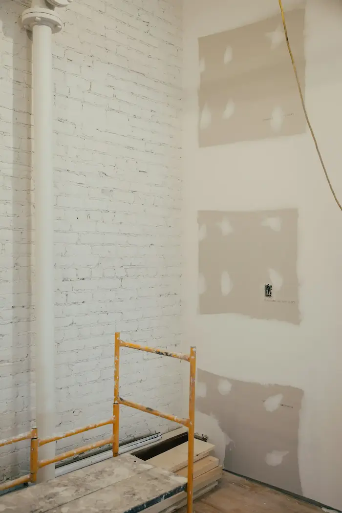 Un mur en briques blanches avec une partie en placo non peinte, une prise électrique visible, un tuyau blanc vertical, et un escabeau en métal jaune au premier plan.