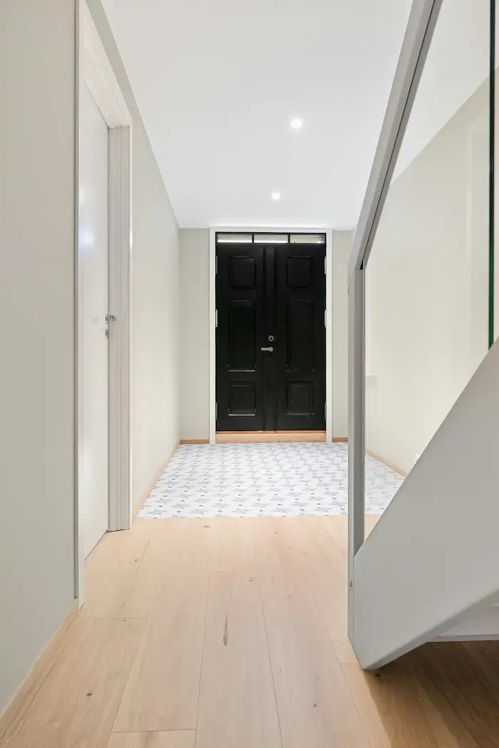 Un couloir avec un sol en bois clair, des murs blancs, une porte blanche à gauche, une porte noire à double battant au fond, et un carrelage à motifs géométriques devant la porte noire.