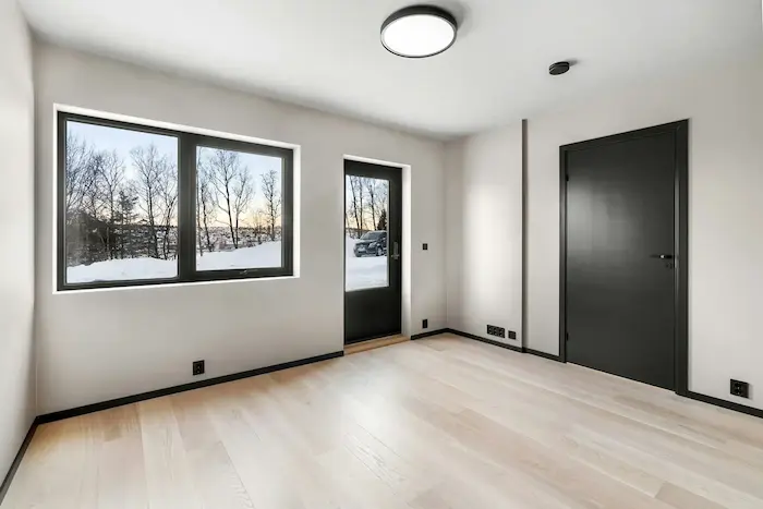 Une pièce vide avec un sol en bois clair, des murs blancs, une grande fenêtre à deux battants sur le mur de gauche, une porte noire sur le mur de droite, et une autre porte vitrée au fond donnant sur un extérieur enneigé avec des arbres dénudés.