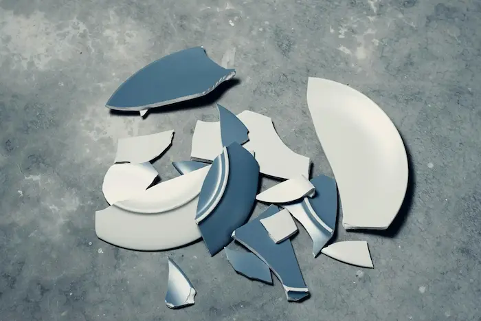 Des morceaux d'assiettes cassées en céramique blanche et bleue dispersés sur un sol en béton.