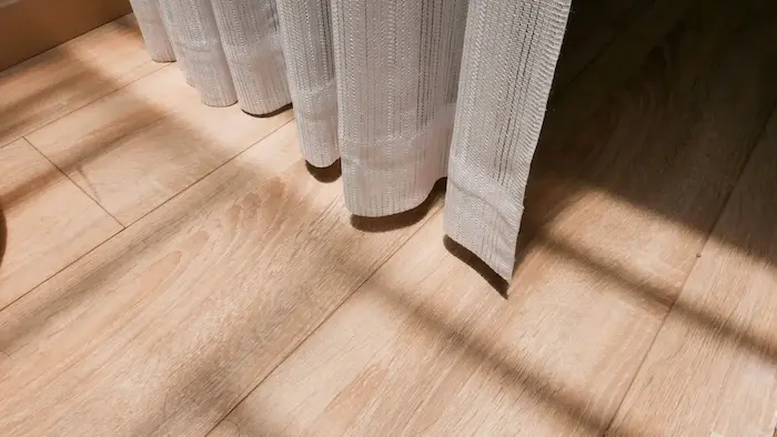 Un sol en bois clair avec des lattes disposées horizontalement, partiellement recouvert par un rideau en tissu blanc texturé, laissant passer des ombres projetées par la lumière du soleil.