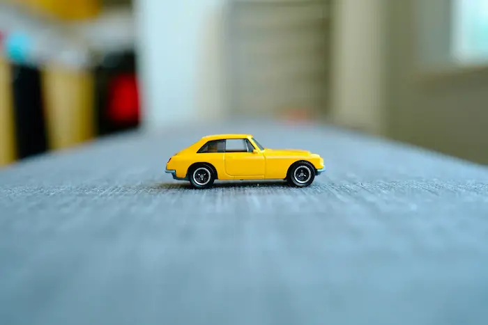 Une petite voiture jaune miniature posée sur une surface grise, avec un arrière-plan flou montrant des éléments colorés indistincts.